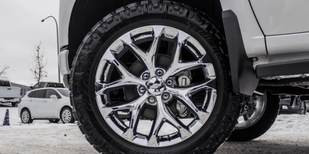 Close-up of a chrome alloy wheel mounted on an SUV, showcasing its sleek design and robust tread pattern suitable for winter driving, with a snowy backdrop and other vehicles in the background.