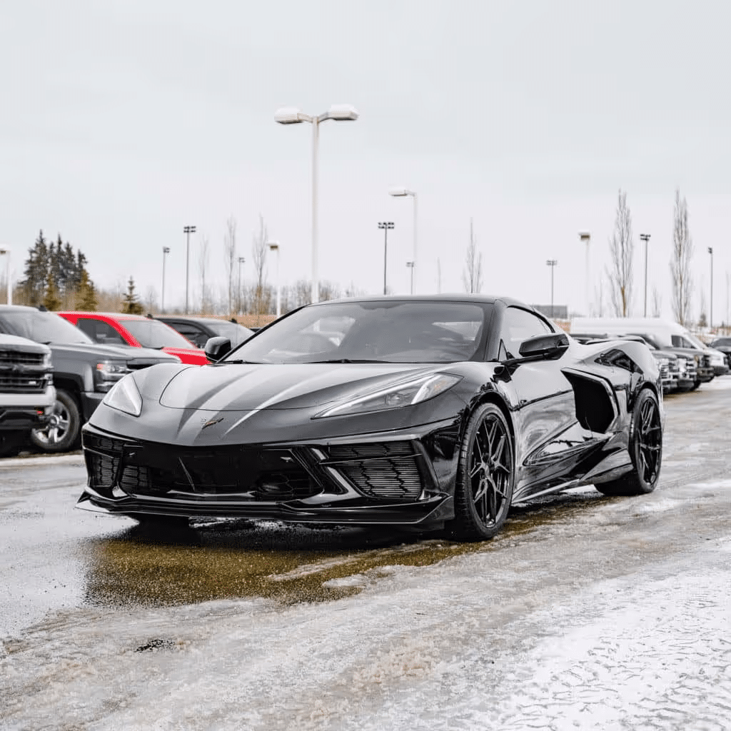 Black Chevrolet Corvette C8 parked in a dealership lot with a row of vehicles in the background, showcasing its sleek design and aggressive stance. The scene is set on a cloudy day with some snow on the ground.