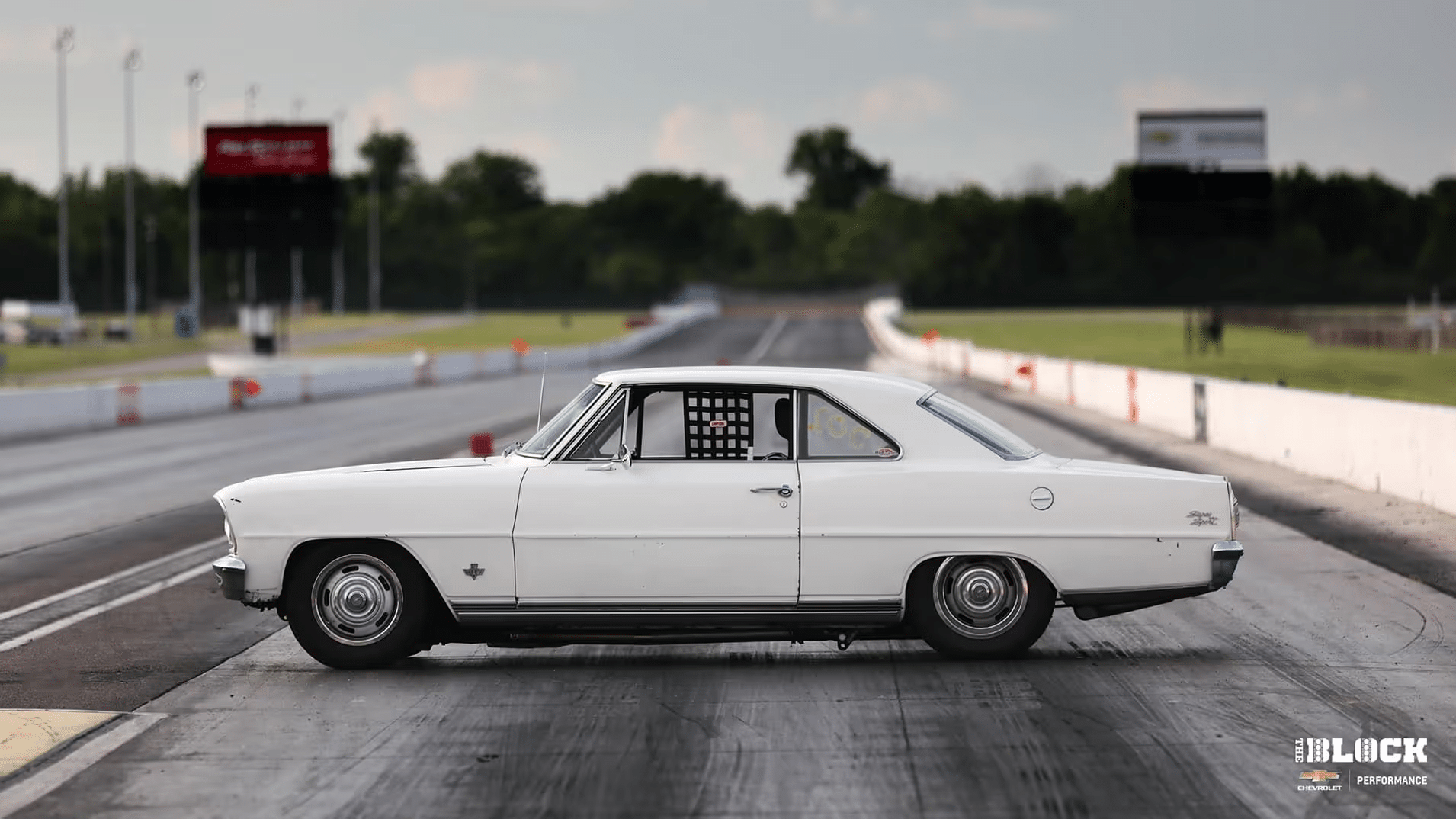 Classic white Chevrolet Nova drag racing car positioned on a racetrack, featuring a roll cage and racing wheels, with an expansive racetrack and trees in the background. Ideal for automotive enthusiasts and drag racing fans.
