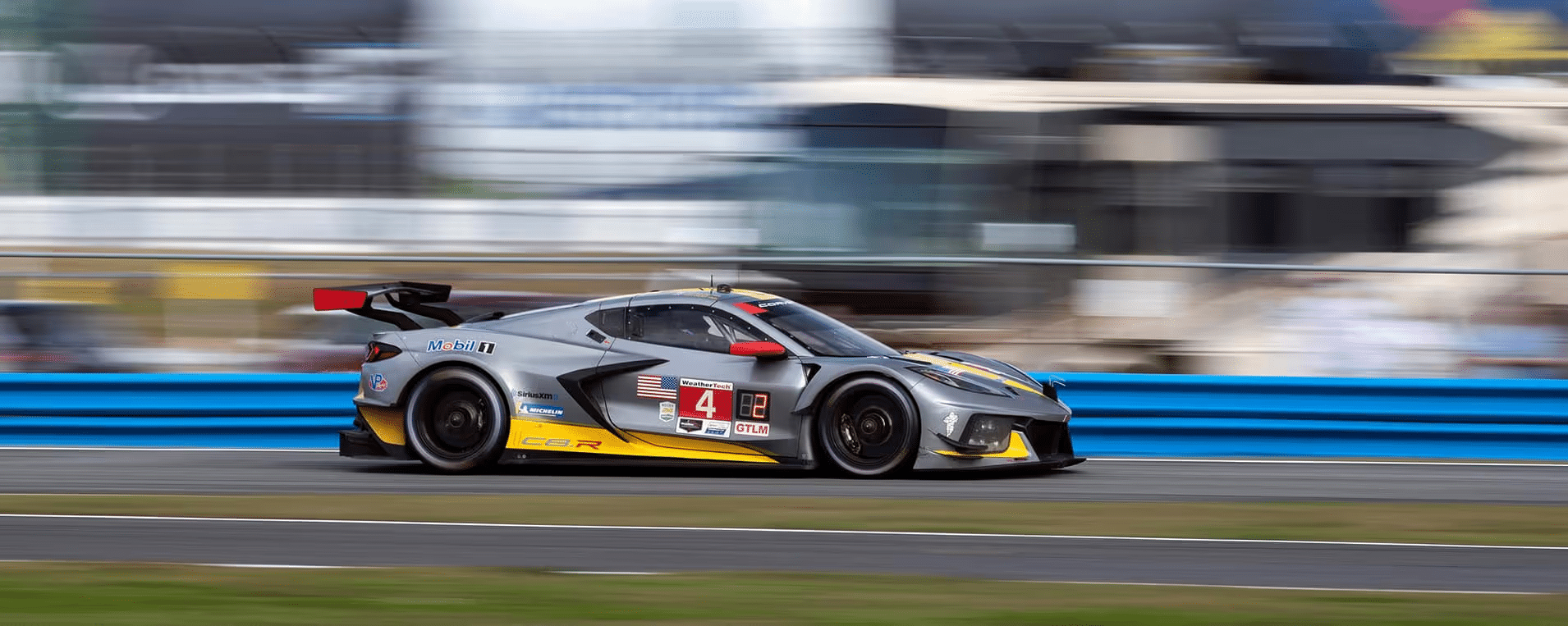 Silver and yellow Chevrolet Corvette C8.R race car speeding on a track during a motorsport event, showcasing a dynamic motion blur effect with a blue barrier in the background.