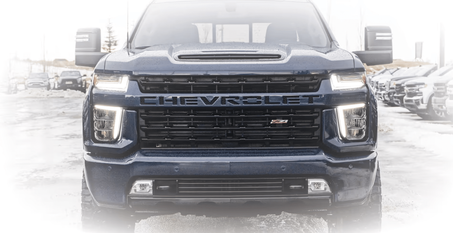 Front view of a rugged black Chevrolet truck featuring off-road modifications, including an aggressive bumper, LED light bar, and large tires, parked in a snowy dealership lot.
