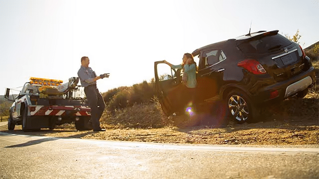 A roadside assistance worker helps a woman who has exited her black SUV after a breakdown. A tow truck is parked nearby under bright sunlight, showcasing a typical roadside emergency scenario.