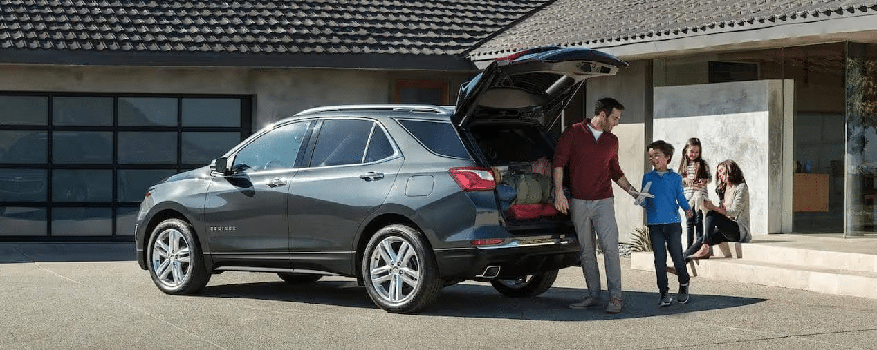 Family enjoying a day out with a gray SUV parked in a driveway, featuring an open trunk filled with luggage, while parents and children prepare for their adventure.