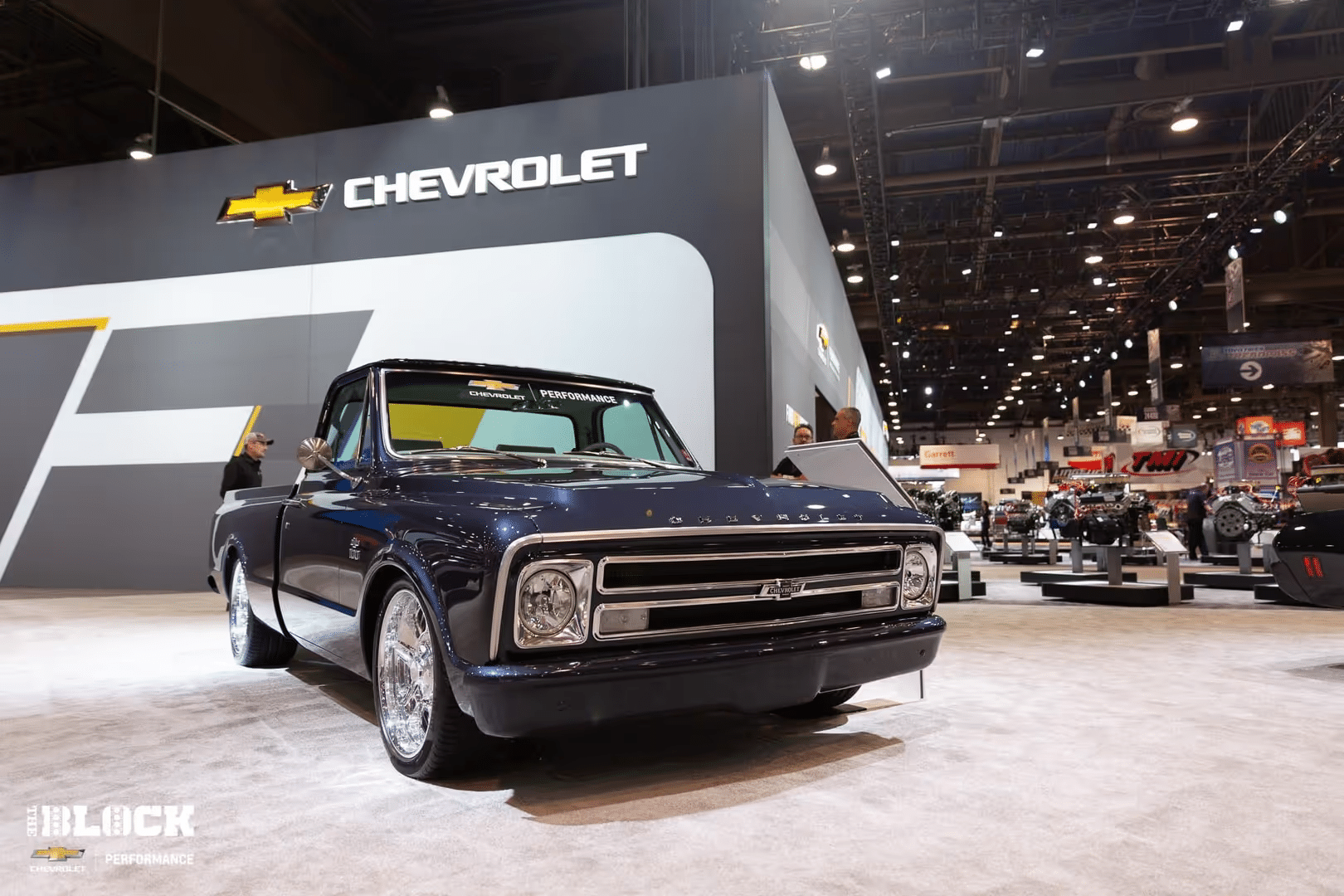 Classic Chevrolet truck displayed at an auto show, featuring a sleek blue exterior and shiny chrome wheels, with the Chevrolet logo prominently featured in the background.