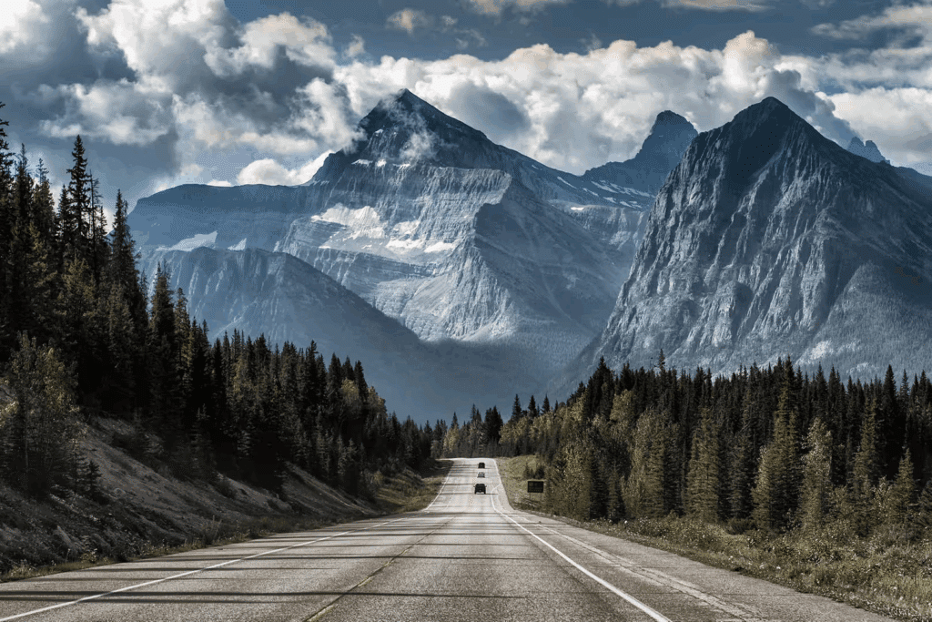 Scenic view of a winding road leading towards towering mountains, surrounded by lush green forests under a dramatic sky filled with clouds. Ideal for travel and nature enthusiasts.