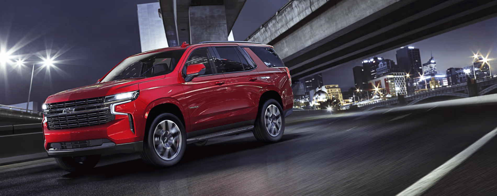 A vibrant red Chevrolet Tahoe SUV is captured in a dynamic nighttime urban setting, showcasing its sleek design and powerful stance as it drives along a lit highway with city lights in the background.