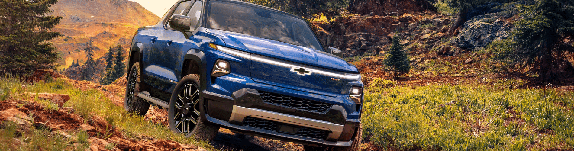 Blue Chevrolet truck driving through rugged terrain with a mountainous backdrop and greenery, showcasing its off-road capabilities and modern design.