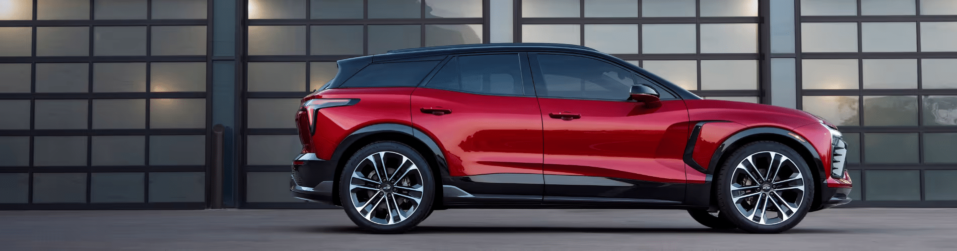 Sleek red SUV parked in front of a modern garage with large glass doors, showcasing an aerodynamic design and stylish alloy wheels. Ideal for showcasing innovative automotive design and performance features.