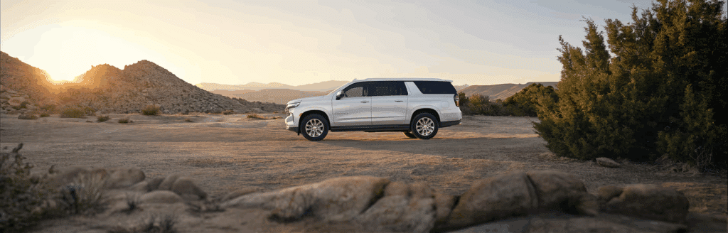 A sleek white SUV parked on an open desert landscape during sunset, surrounded by rocky terrain and sparse vegetation. The warm sun casts a glow over the scene, highlighting the vehicle's modern design and features.