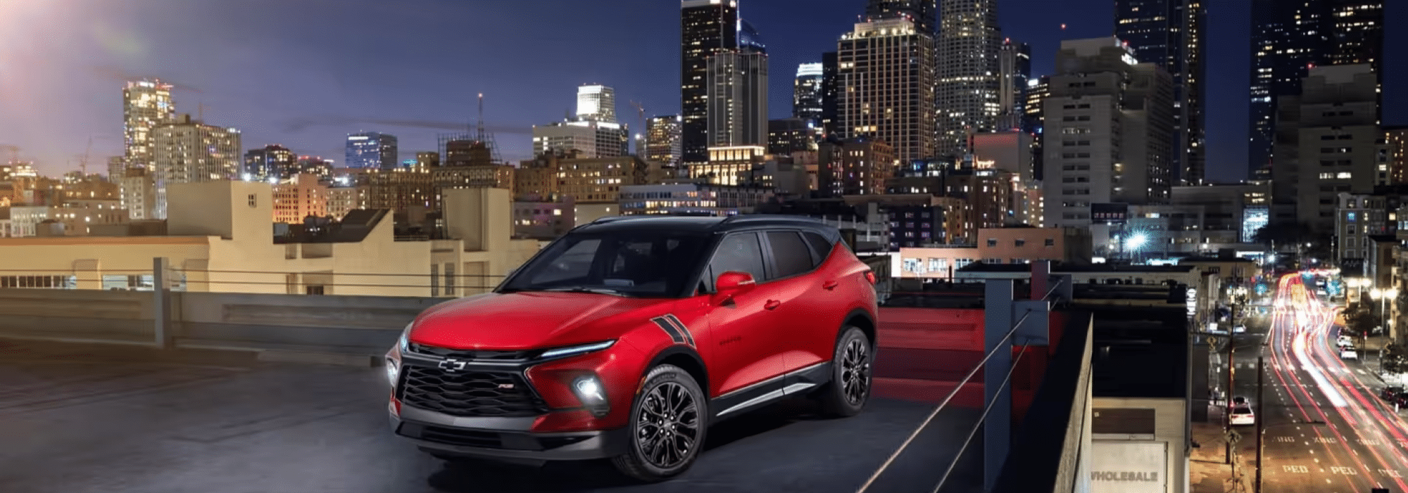Chevrolet Blazer SUV parked on a rooftop with a city skyline backdrop at night, highlighting its bold red color and stylish design against illuminated buildings.