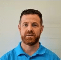 Portrait of a man with a beard wearing a blue polo shirt, standing against a light background, showcasing a neutral expression.