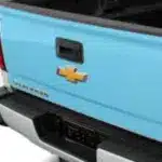 Close-up of the rear tailgate of a light blue Chevrolet truck, featuring the iconic Chevy bowtie emblem and tailgate latch.