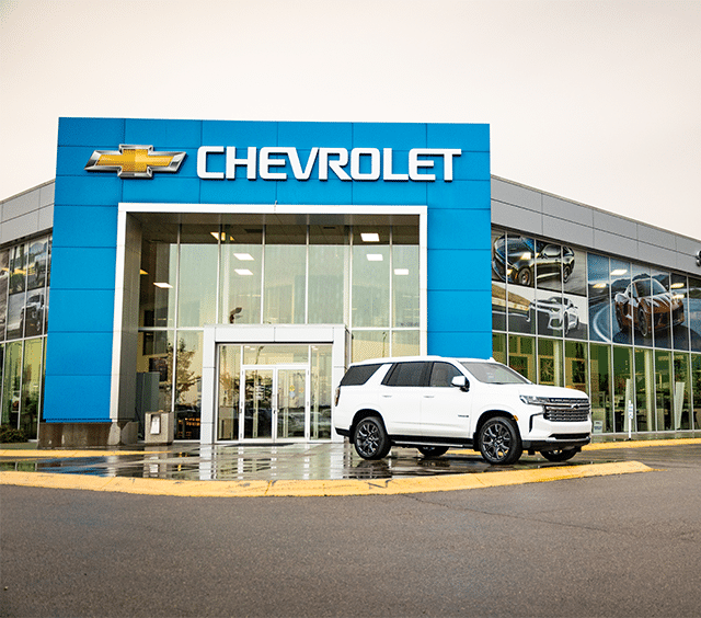 Chevrolet dealership exterior featuring a new white Chevrolet SUV parked in front of the modern building with a blue Chevrolet sign, showcasing promotional images of vehicles in the windows.