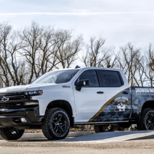 White Chevrolet Silverado truck parked on a gravel road, featuring a black graphics wrap promoting Sherwood Park and Rocky Ridge. Trees in the background under a clear sky.