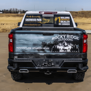 Rear view of a Chevrolet truck featuring a Rocky Ridge Outdoors graphic and promotional decals for Sherwood Park Chevrolet, highlighting its status as the #1 volume dealer in Canada. The background showcases a scenic landscape ideal for outdoor adventures.