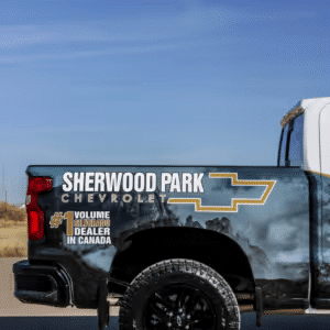 Side view of a pickup truck featuring the Sherwood Park Chevrolet logo, showcasing its title as the number one volume dealer in Canada. The truck is parked on a scenic road with a clear blue sky in the background, emphasizing its rugged design and branding.
