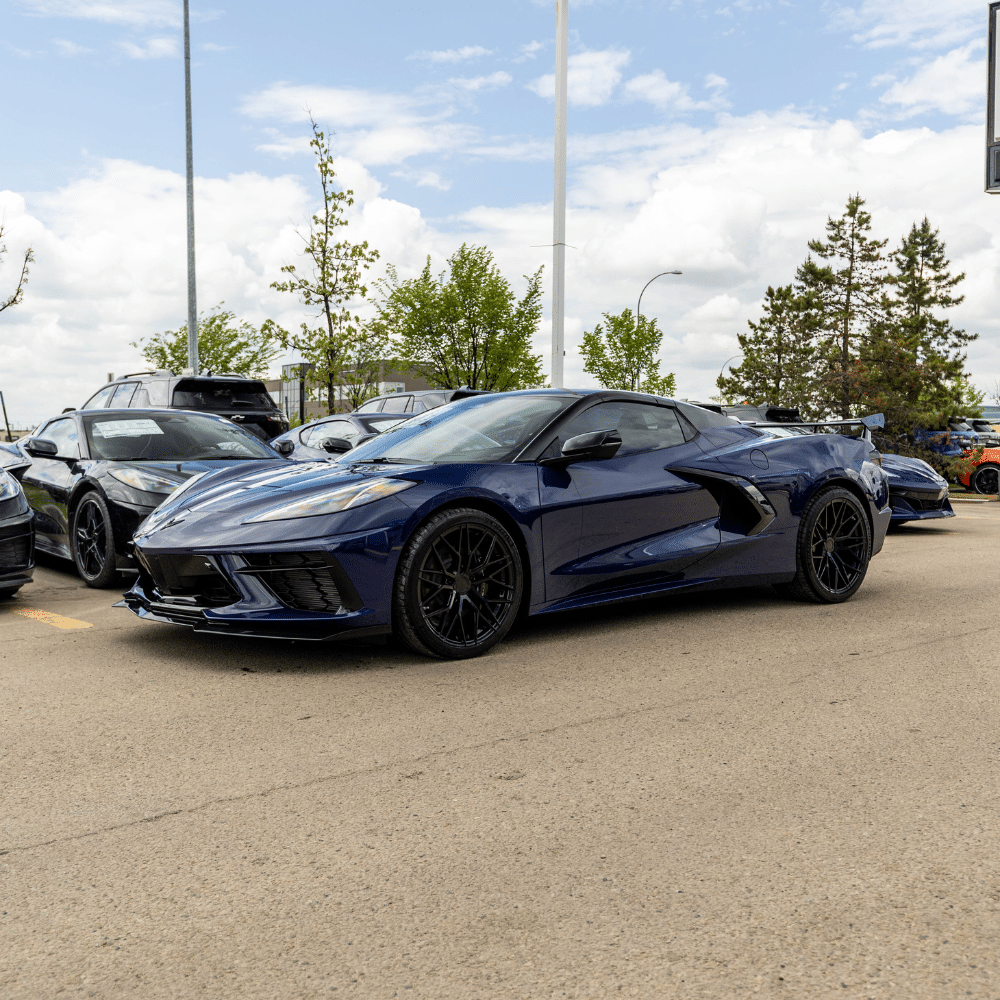 Blue sports car parked outdoors under a cloudy sky, surrounded by other vehicles.