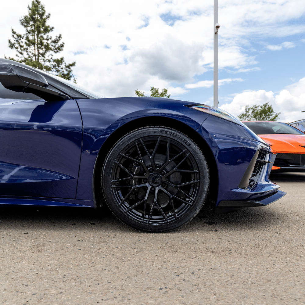 Close-up of a blue sports car wheel with black rims in a parking lot, featuring another car in the background.