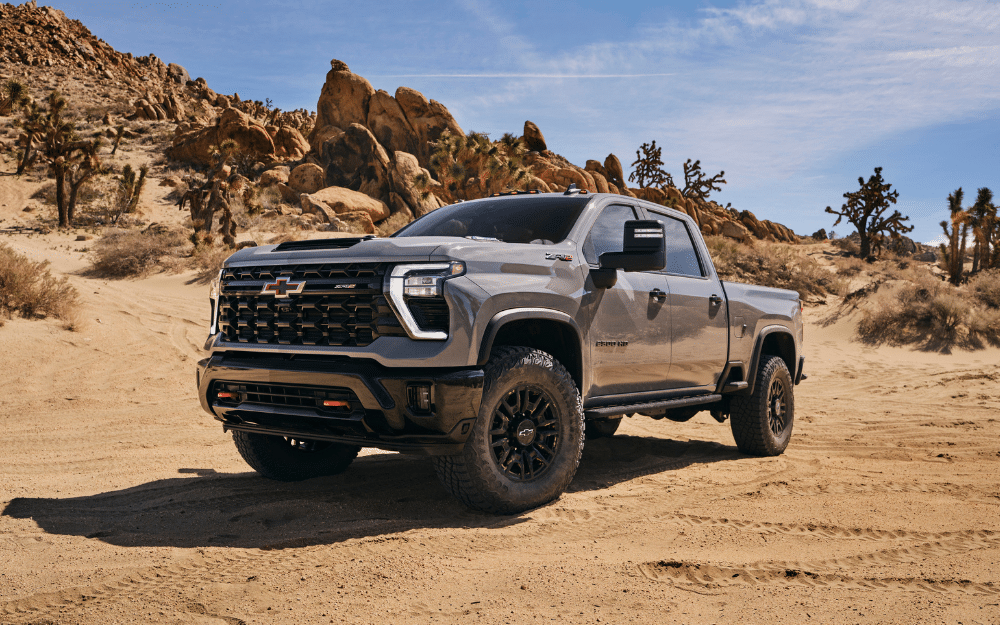 A rugged pickup truck on a sandy desert terrain with rocky formations and clear blue sky in the background.