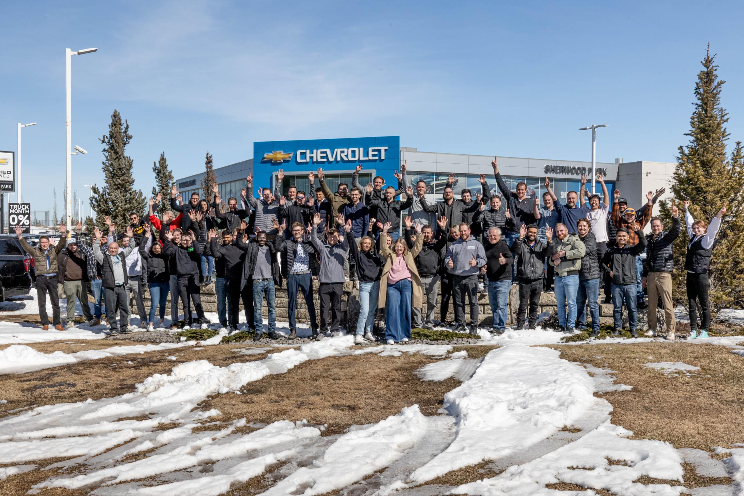 A large group of people posing and cheering outside a Chevrolet dealership, with snow on the ground.